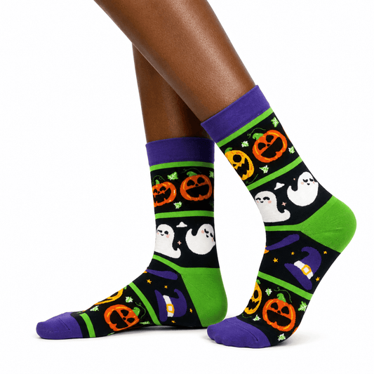 A pair of crew socks with a Halloween pattern, shown on a person's feet against a plain white background. The socks feature a black background with green stripes, orange jack-o'-lanterns, white ghosts, purple cuffs and toes, and a purple witch hat motif.