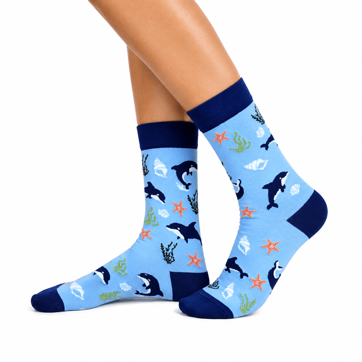 A pair of light blue crew socks with a dark navy blue cuff, heel, and toe, featuring a playful pattern of black and white orcas, orange starfish, and green seaweed, shown on a model's feet against a white background.