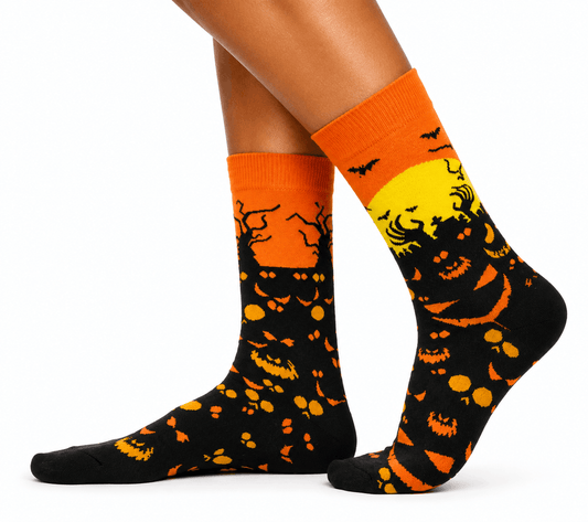 A pair of crew socks worn on a person's feet, featuring a Halloween design with orange and black jack-o'-lantern patterns and a yellow full moon on a black background.