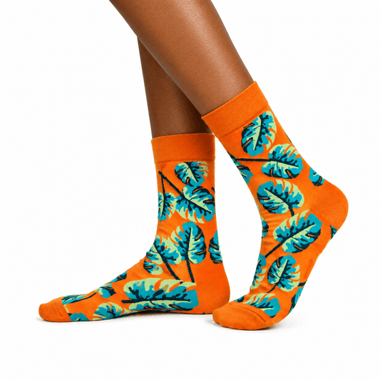 A pair of crew socks with a bright orange base and a teal and yellow tropical leaf pattern, shown on a model's feet against a white background.