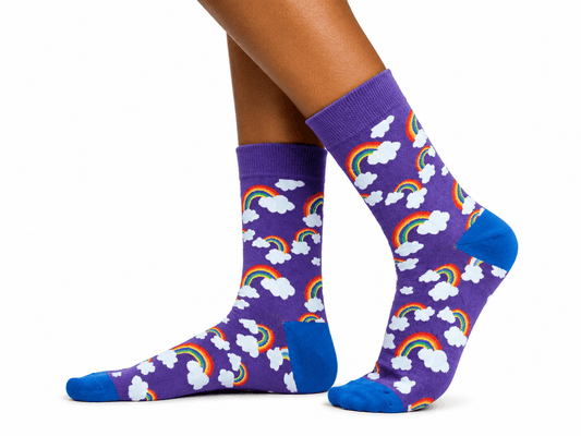 A pair of purple crew socks with a repeating pattern of white clouds and colorful rainbows, worn on a person's feet against a plain white background.