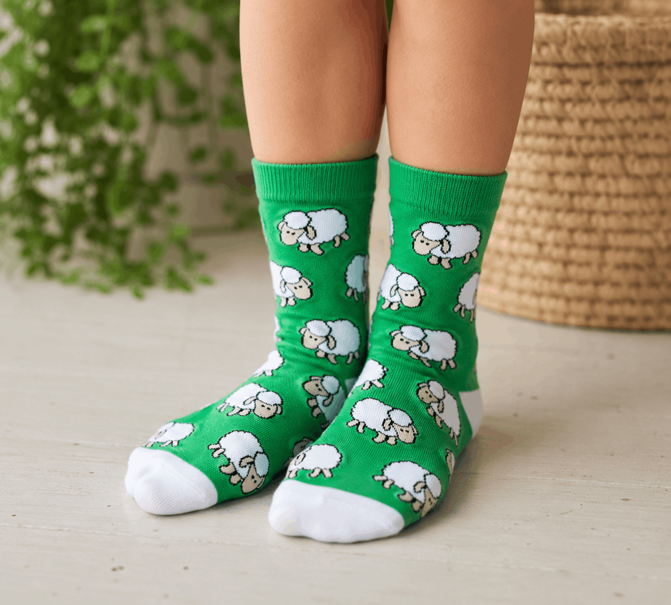 A pair of bright green crew socks with a repeating pattern of white sheep, worn on a child's feet. The background features a light-colored floor, a woven basket, and a green plant.