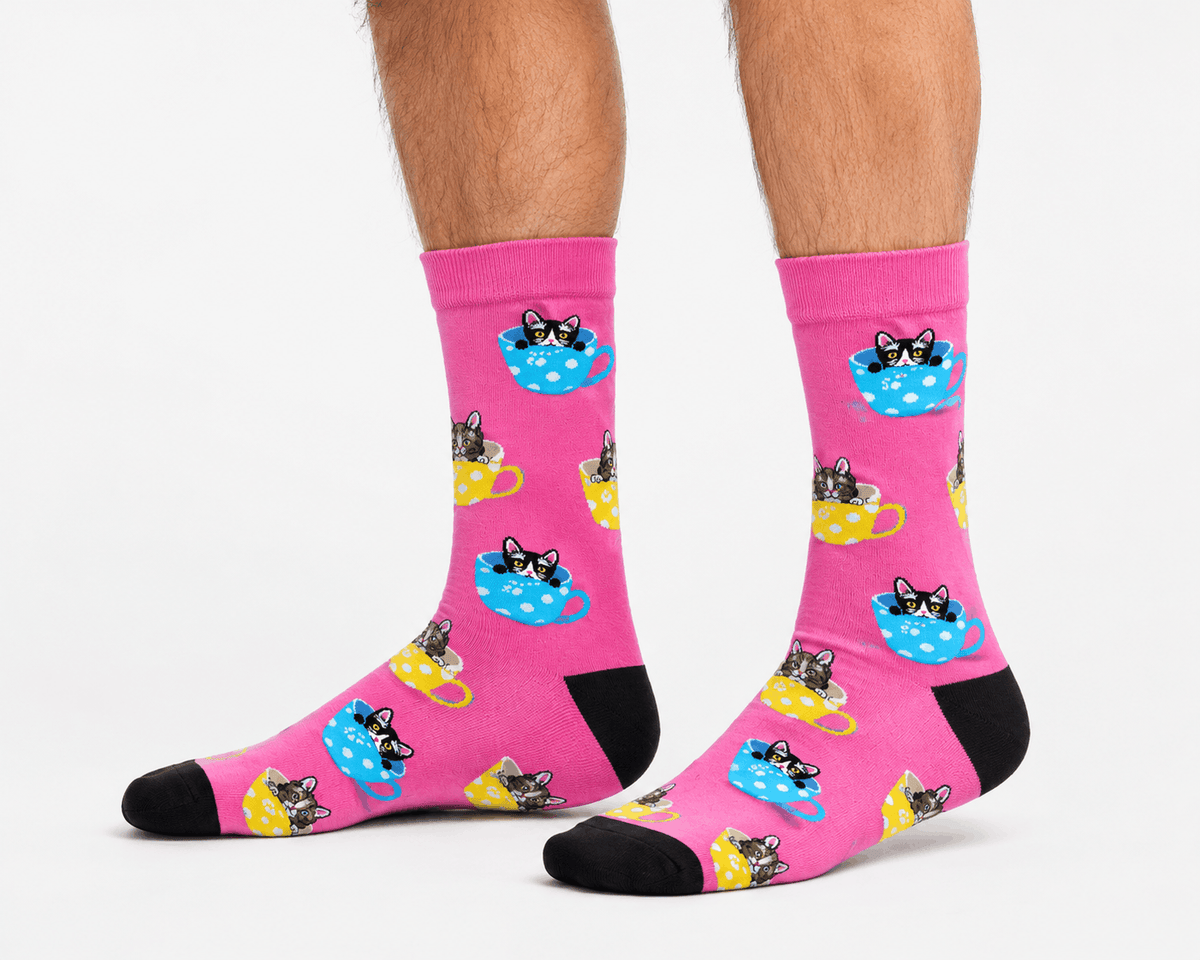 A pair of bright pink crew socks with a repeating pattern of cartoon cats lounging in teacups, worn on a person's feet against a clean white background.